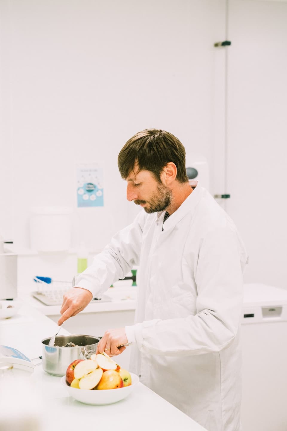 A technician in a white coat cooks fruit in a saucepan, next to a bowl of sliced apples