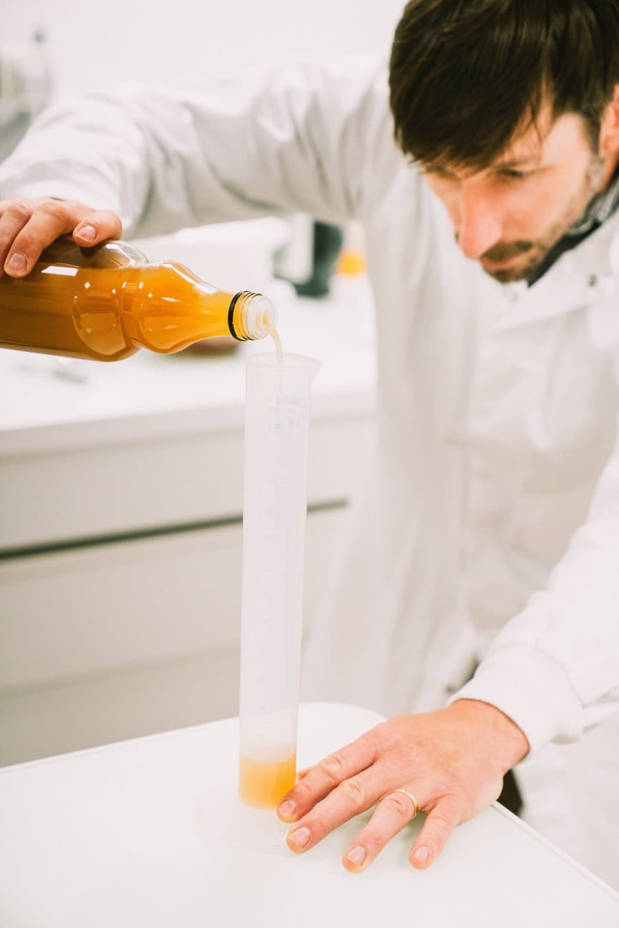A technician in a white coat pours apple juice into a graduated cylinder to analyse it